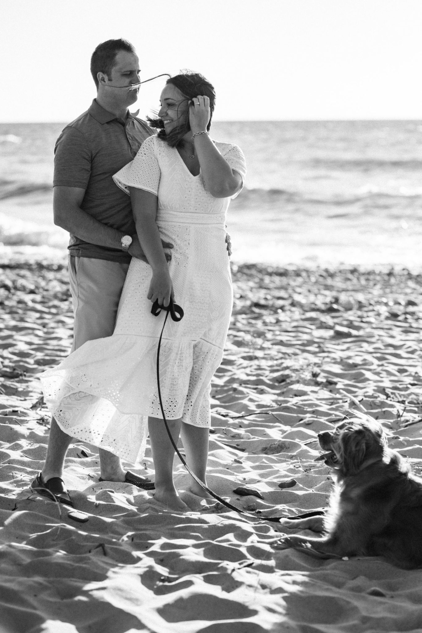 Black and white engagement photo of a couple holding their dog on the beach.