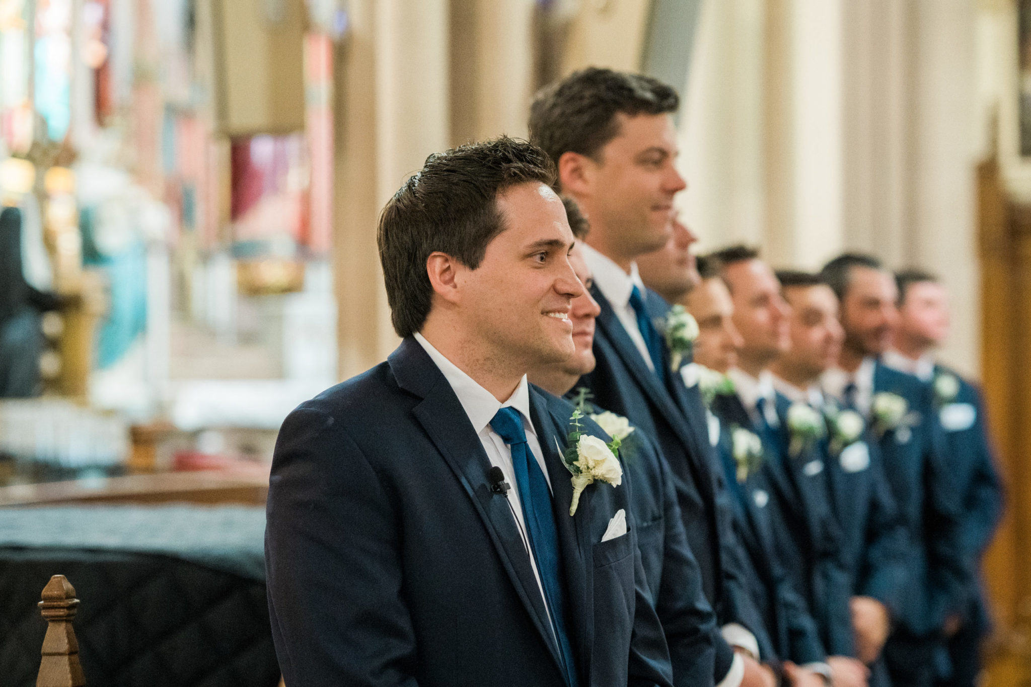 a groom standing at the end of the aisle in the basilica of sainte anne de detroit and smiling at his bride