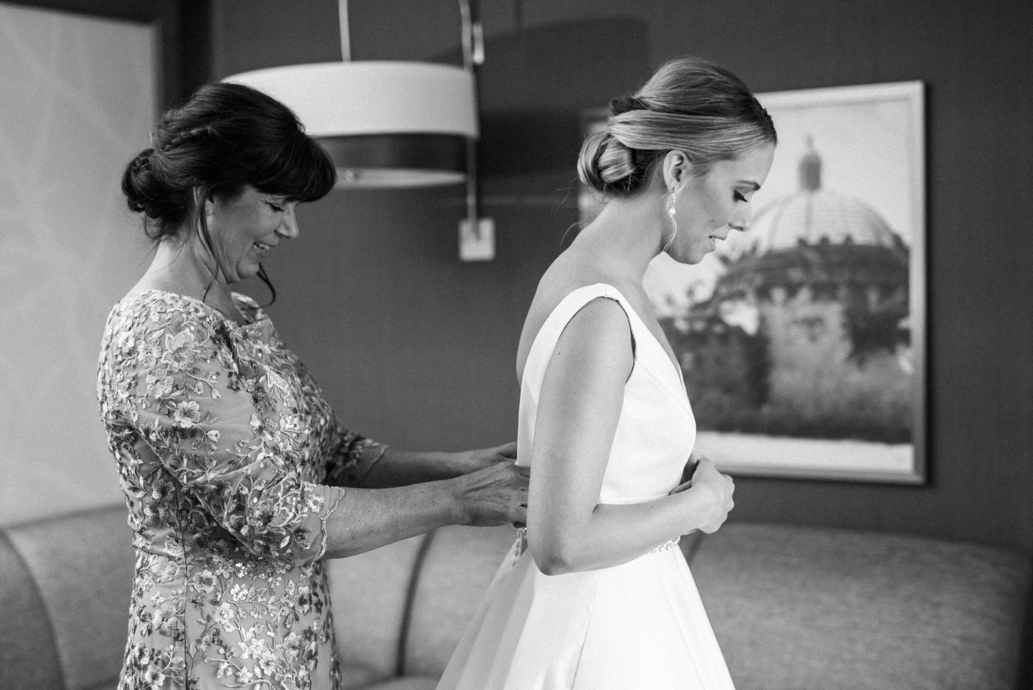 a black and white image of a mother helping her daughter into her classic wedding dress