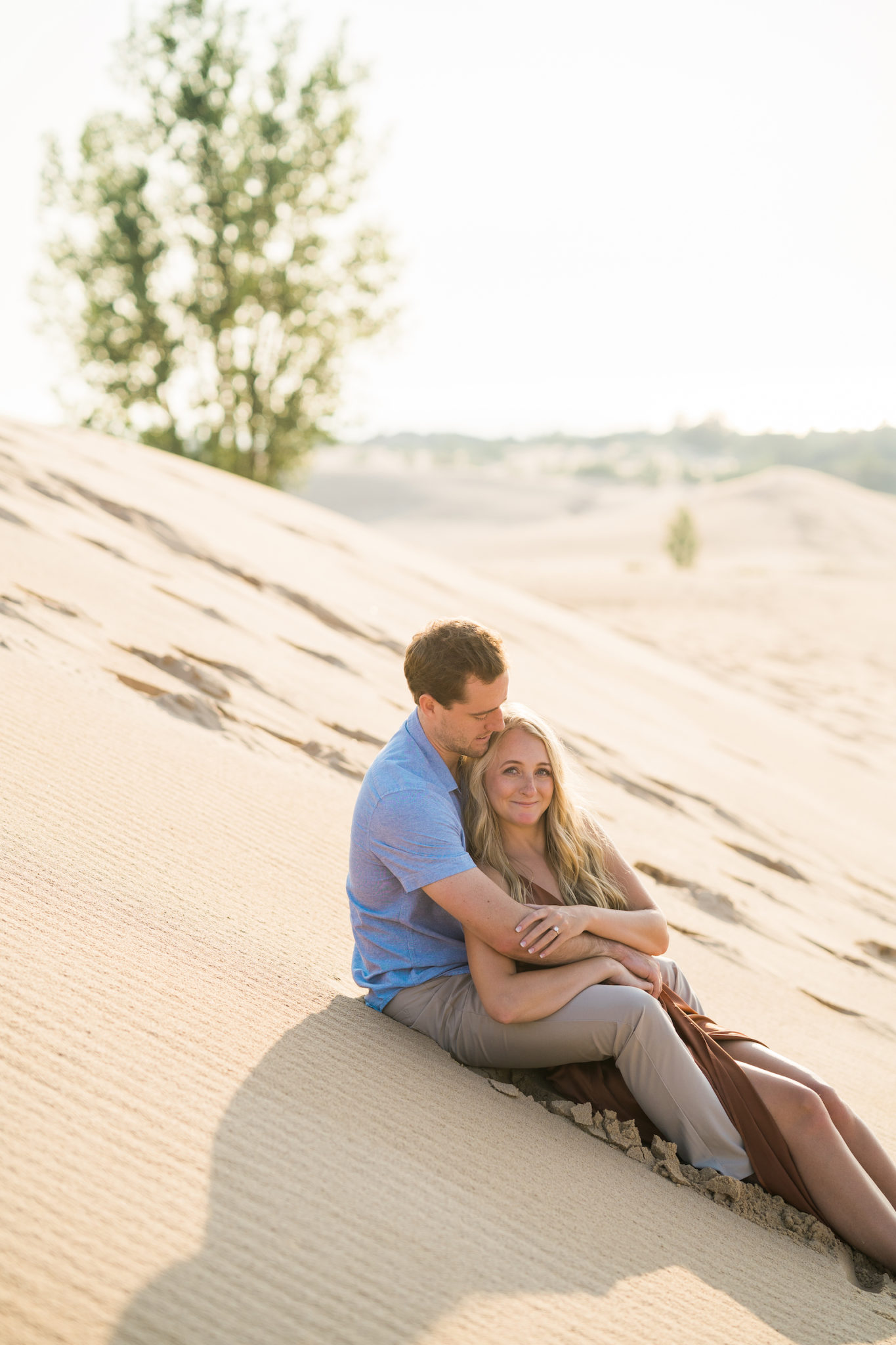 a couple sitting on the sand dunes and holding one another during their summer engagement photos