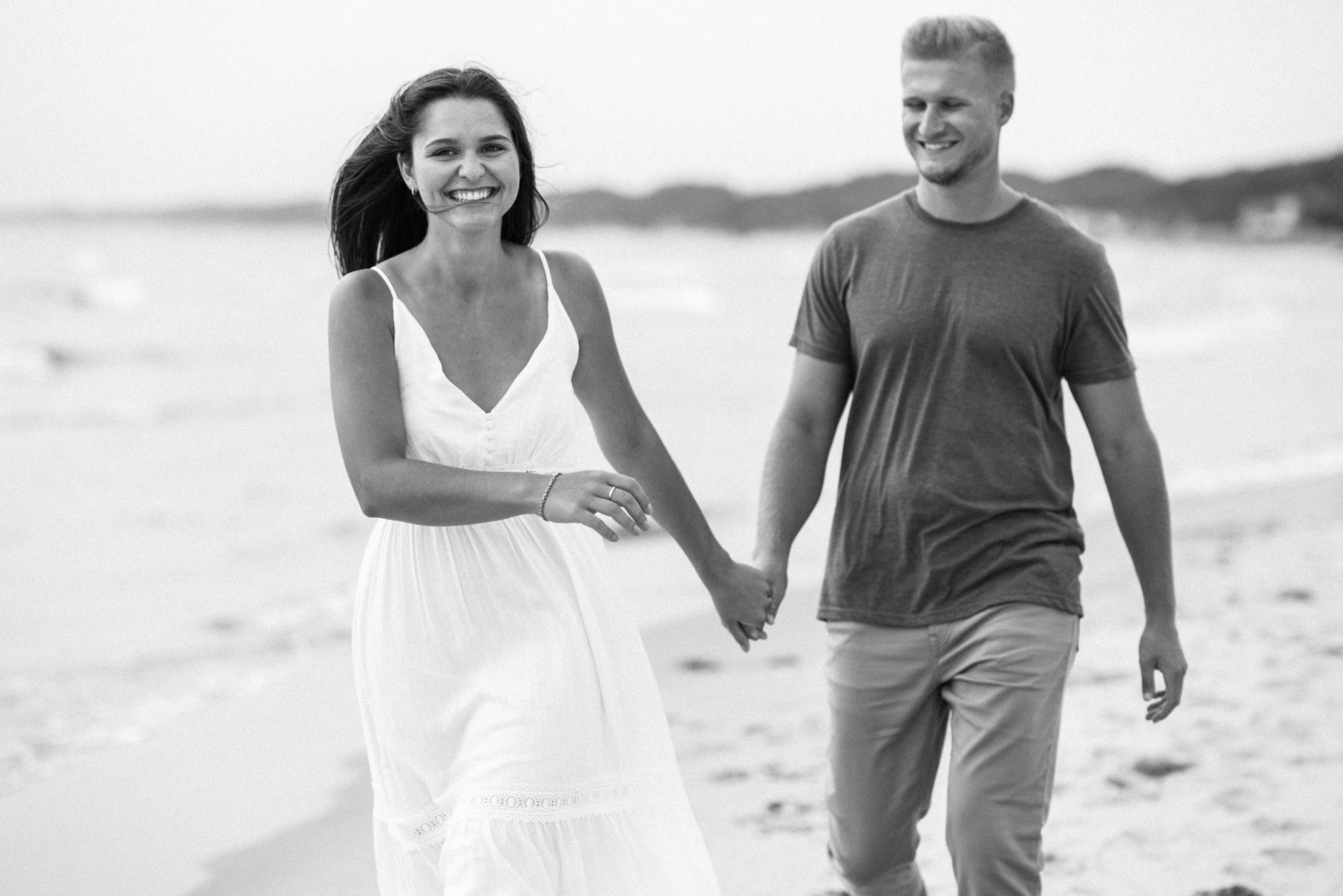 a black and white image of a couple holding hands and walking during their grand haven beach engagement photo session in michigan